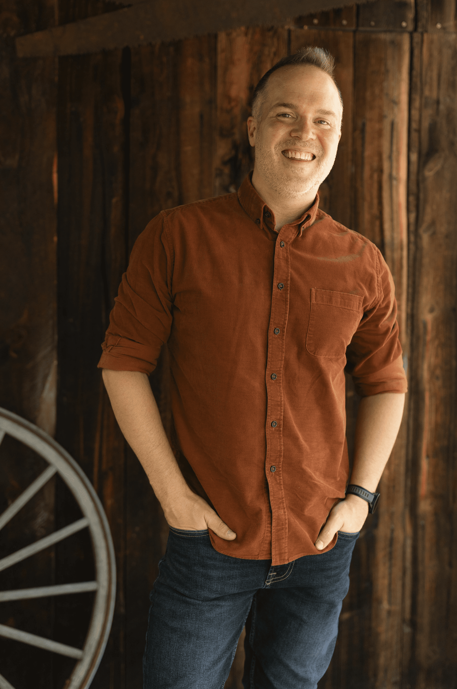 Man in a rust-colored button-down shirt standing in front of a wooden wall with a wagon wheel visible on the left.